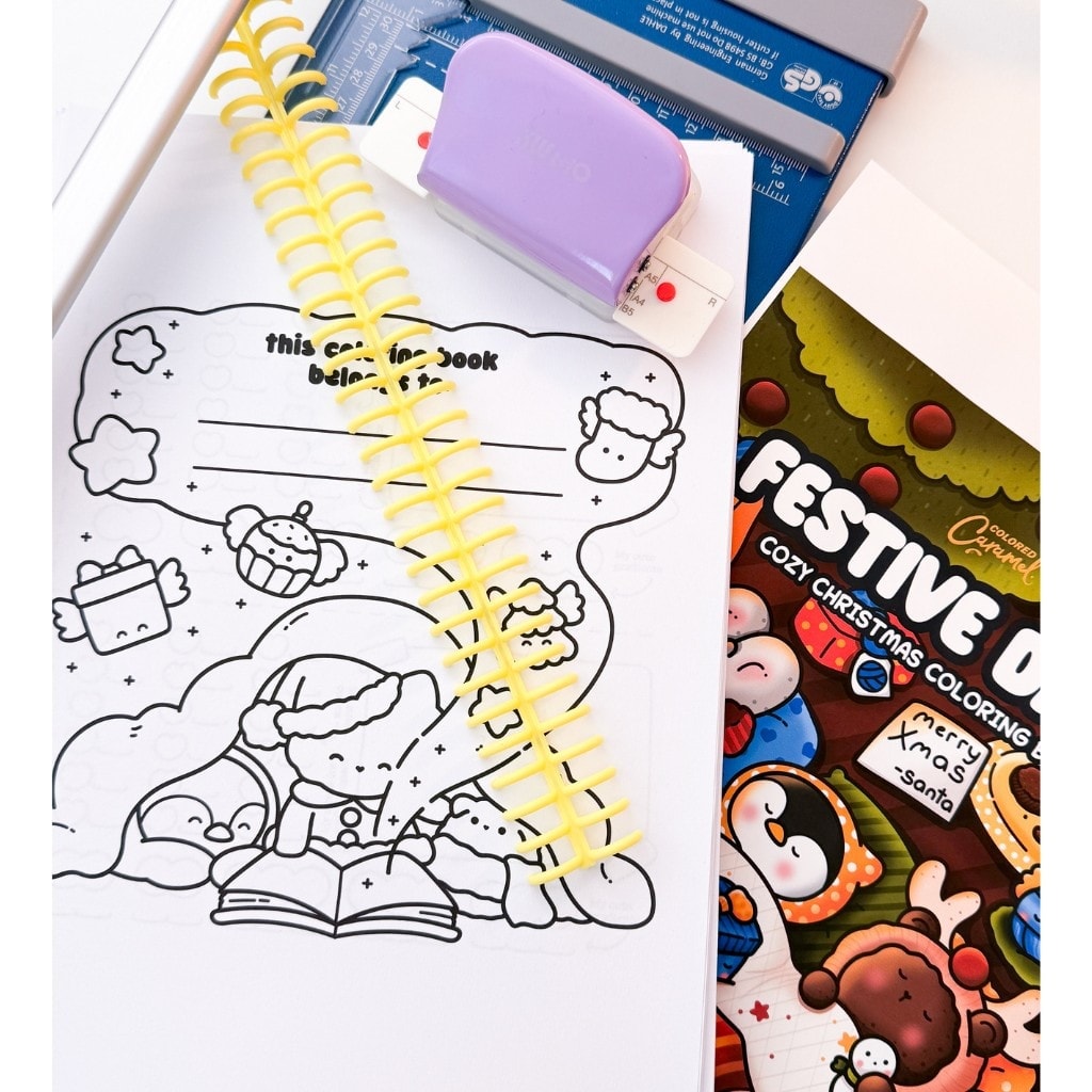 Spiral binding coil, hole puncher, paper cutter, and printed coloring pages laid out for assembling a spiral-bound coloring book.