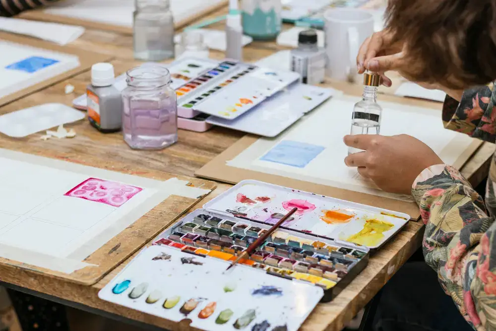 Künstlerin malt Aquarell auf Papier mit Palette und Pinseln auf einer hölzernen Arbeitsfläche.