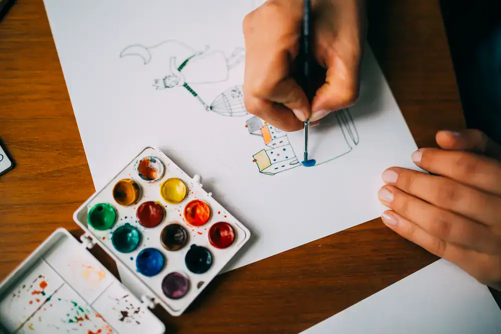 Handmalen einer kleinen Illustration mit Aquarellfarbe und Pinsel neben einer Aquarellpalette auf einem Blatt Papier.