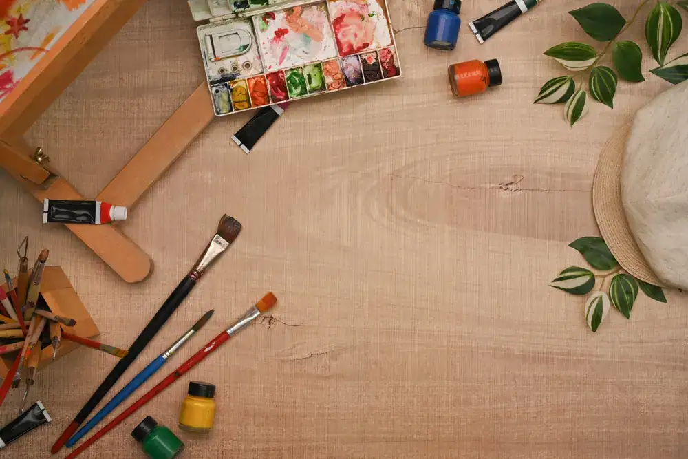 Aquarellmalutensilien auf einem Holztisch mit Pinseln, Aquarellpalette, Farbtuben und kleinen Farbgläsern.