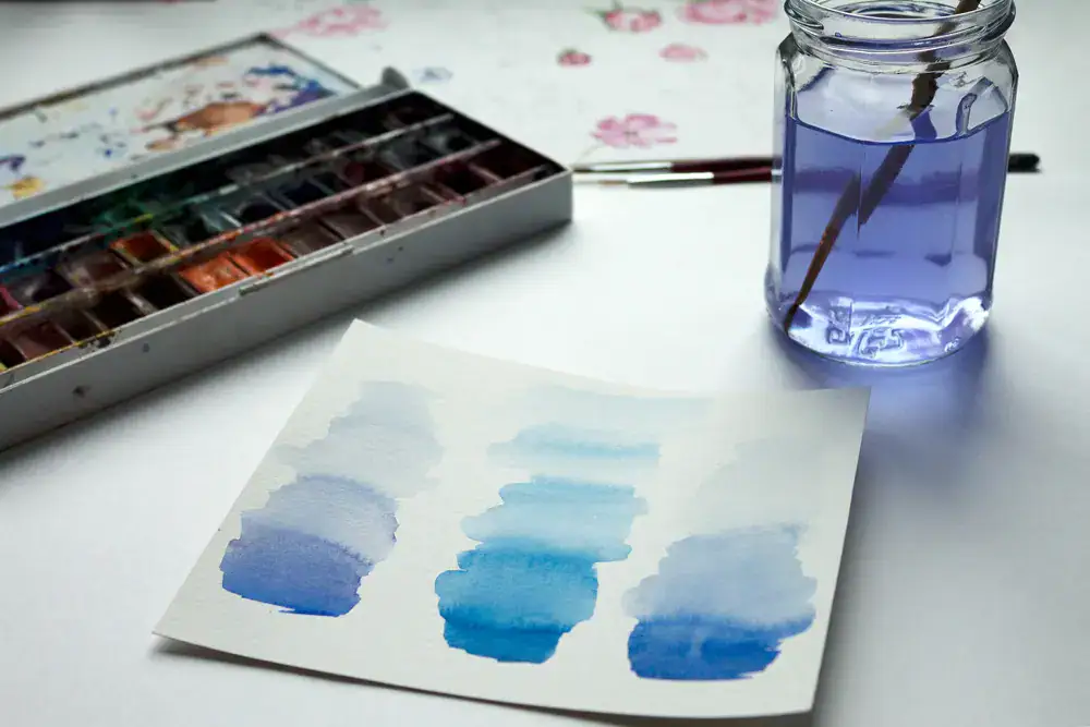 Aquarelle mit Farbverläufen auf Papier neben einer Palette und einem Pinsel in einem Glas mit Wasser.