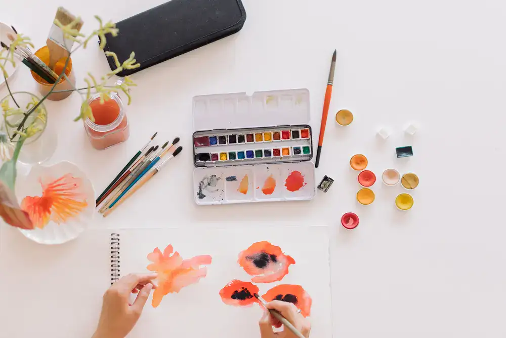 Künstlerin malt Aquarellblumen in einem Skizzenbuch mit Palette und Pinseln auf einer sauberen Arbeitsfläche.